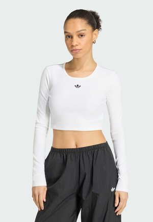 Mlada žena s kovrčavim repom nosi bijelu majicu s dugim rukavima i široke crne hlače, obje s malim Adidas logom, stoji na jednobojnoj pozadini.