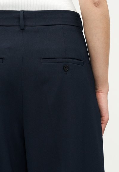 Κοντινό πλάνο σκούρων μπλε παντελονιών με πίσω τσέπη με κουμπί, φορεμένα με λευκό τοπ, που δείχνει μέρος του βραχίονα του φορέα.