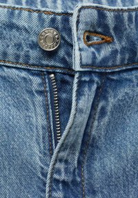 Vaaleansiniset farkut, joissa on metallipainike, vetoketjukiinnitys ja kontrastivärinen ruskea ommel, jossa on hieman kuluneen näköinen pinta.