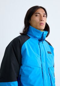 Blaue und schwarze wasserdichte Jacke mit hohem Kragen, Reißverschluss und Logo-Patch; bietet strukturierten Stoff und kontrastierende Ärmel.