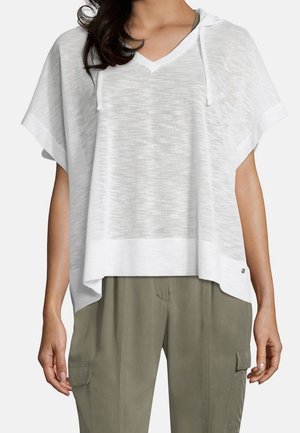 Mujer con una blusa blanca, holgada y de manga corta con capucha, sobre pantalones cargo verde oliva, de pie con los brazos relajados a los lados.