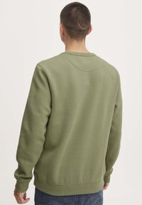 Uomo con capelli corti e una cicatrice curva visibile sulla parte posteriore della testa, indossa una felpa verde oliva e jeans blu, di spalle.