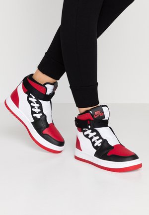 Osoba w czarnych spodniach i czerwono-biało-czarnych sneakersach Nike Air typu high-top, stojąca na białej powierzchni.