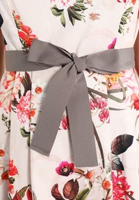 Abito floreale con fiori rosa, rossi e arancioni, legato in vita con una larga cintura di tessuto grigio a forma di fiocco.