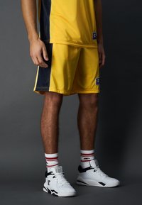 Pantalones cortos deportivos amarillos con paneles laterales en navy, combinados con zapatillas blancas con detalles en negro y calcetines a rayas rojas. Tejido suave, corte relajado.