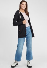 Svart hettejakke med glidelås, med kremfargede snorer og trekantede brune detaljer. Kombinert med lyseblå vide jeans og hvite støvler.