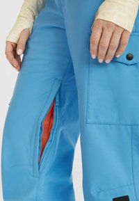 Blauwe sneeuwbroek met een gladde textuur, met een net gevoerde pocketopening in oranje en een zijzak met een zwarte drukknoopsluiting.
