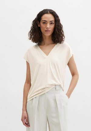 Vrouw met krullend haar die een beige V-hals top met korte mouwen en lichtgrijze hooggetailleerde geplooide broek draagt, staande met één hand in de zak.