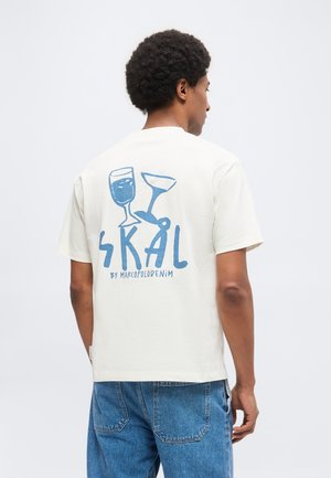 Hombre con camiseta blanca con gráfico azul de dos vasos para beber y la palabra "SKÅL" en la parte trasera, combinado con jeans azules.