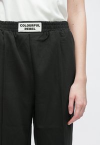 Melnas, vaļīgas bikses ar elastīgu jostu. Aizmugurē ir balta etiķete ar uzrakstu "COLOURFUL REBEL." Gluda tekstūra.