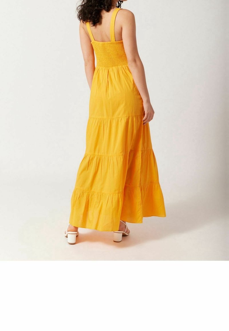 Robe maxi jaune avec un corsage smocké, présentant des panneaux de tissu superposés et des bretelles réglables. Portée avec des sandales blanches à talons.