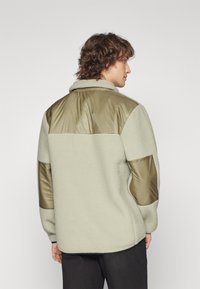Giacca in pile di un beige chiaro con accenti in nylon verde oliva sulle spalle e sulle maniche. Presenta un colletto e un tessuto testurizzato.