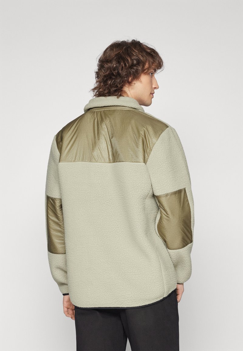 Fleecejacka i ljusbeige med olivgröna nylonaccent på axlar och ärmar. Har en krage och strukturerad tyg.