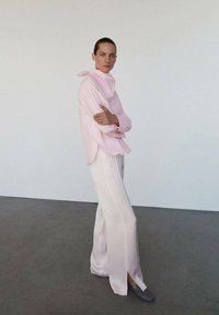 Chemisier rose clair, ample, avec un col haut et un détail écharpe, associé à un pantalon large et fluide blanc avec des fentes latérales et des chaussures grises.