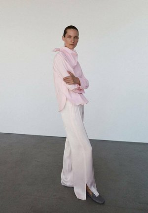 Chemisier rose clair, ample, avec un col haut et un détail écharpe, associé à un pantalon large et fluide blanc avec des fentes latérales et des chaussures grises.