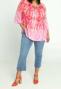 Donna che indossa una blusa larga a fantasia rosa e rossa, jeans blu a vita bassa, e sandali con tacco arancioni brillanti, in piedi con una mano in tasca.
