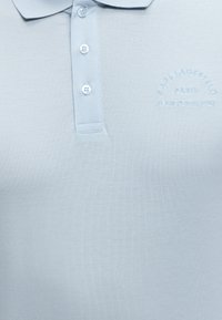 Ανοιχτό μπλε πόλο μπλουζάκι με τρία κουμπιά και κεντημένο κείμενο "Karl Lagerfeld Paris 21 Rue St-Guillaume" στο στήθος.