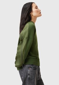 Grön sweatshirt med en slät textur, med dragkedja på sidan, ribbad nederkant och en avslappnad passform. Matcher med högmidjade jeans.