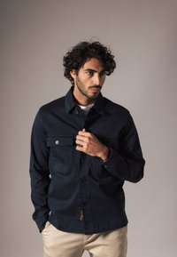 Navyblauwe jas met een knoopsluiting aan de voorkant, twee borstzakken en een gladde stoftextuur. Kenmerkende stikseldetails aan de zoom.