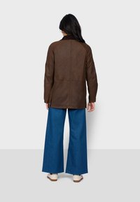 Brązowa skórzana kurtka z zamszowym kołnierzem, o dopasowanym kroju, z pionowymi szwami i bocznymi guzikami. W połączeniu z szerokimi niebieskimi dżinsami.