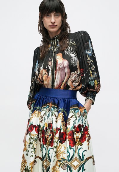 Schwarze Bluse mit künstlerischem Druck historischer Persönlichkeiten; kombiniert mit einem blauen Taillenband und einem Rock mit Blumendruck in Creme, Rot und Grün.