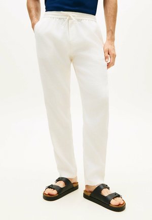 Mężczyzna w białych spodniach ze sznurkiem i czarnych sandałach, stojący z jedną ręką w kieszeni na jednolitym tle.