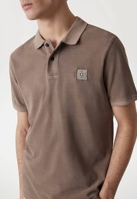 Ruda medvilnės polo marškinėliai su tekstūriniais paviršiais, trijų mygtukų apykakle ir logotipo ženkleliu kairiajame krūtinės šone. Trumpomis rankovėmis ir laisvu siluetu.