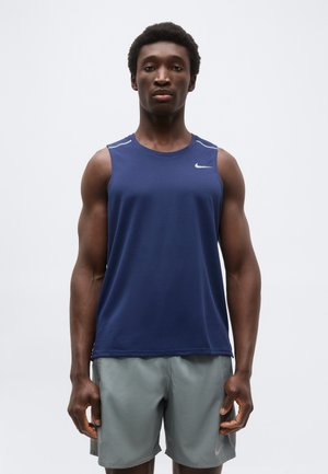 Atletický muž stojaci čelom vpred, oblečený v tmavomodrom tielku Nike bez rukávov a svetlosivých kraťasoch na jednofarebnom pozadí.