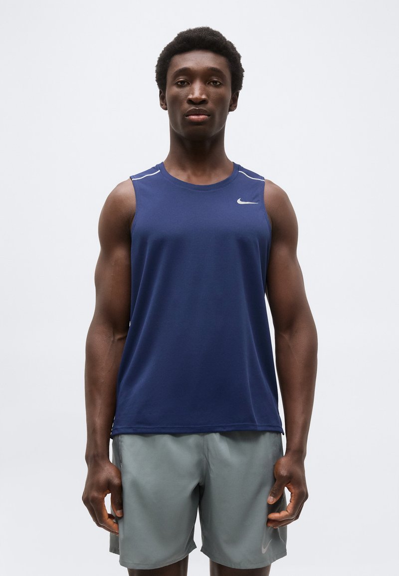 Αθλητικός άντρας που στέκεται με το πρόσωπο προς τα εμπρός, φορώντας μπλε αμάνικη μπλούζα Nike και ανοιχτό γκρι σορτς, μπροστά σε απλό φόντο.