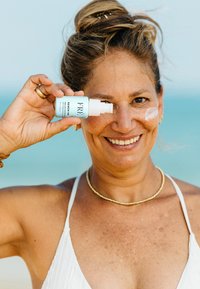 Jasnoniebieska butelka kosmetyków z napisem "ODŚWIEŻ MNIE", trzymana w górze, z białą smugą kremu na policzku modelki. Tło z słoneczną plażą.