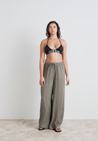 Svart triangulär bikiniöverdel med vitt logotyp, matchad med lösa olivgröna byxor. Slät tygtextur, avslappnad passform och platta sandaler.