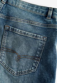 Modré džínsy z denimu s zadnou kapsou s zaobleným dizajnom a kontrastným hnedým prešívaním, ktoré zvýrazňujú textúru a opotrebovanie.