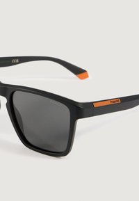 Μαύρα γυαλιά Polaroid με τετράγωνο σκελετό και σκούρους φακούς, με πορτοκαλί λεπτομέρειες στους βραχίονες και τις άκρες τους, σε άσπρο φόντο.