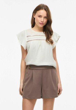 Frau mit langen braunen Haaren, die eine weiße, strukturierte Bluse mit Spitzen-Detail und hoch taillierte braune Shorts mit Knopfakzenten trägt.
