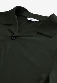 Polo shirt verde scuro realizzato in tessuto testurizzato, con colletto a punta e chiusura con un solo bottone. Etichetta del marchio visibile all'interno del colletto.
