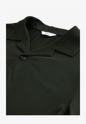 Dunkelgrünes Poloshirt aus strukturiertem Stoff, mit spitzem Kragen und einem einzigen Knopfverschluss. Markenschild im Kragen sichtbar.