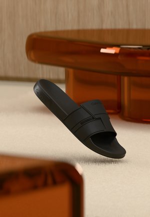 Fekete Calvin Klein papucs a levegőben, bézs színű szőnyeg és barna áttetsző bútorok előtt, egy minimalista szobában.