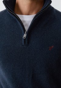 Man i en mörkblå zip-up tröja över en vit crew-neck t-shirt, med en liten broderad röd fågel-logotyp på bröstet.
