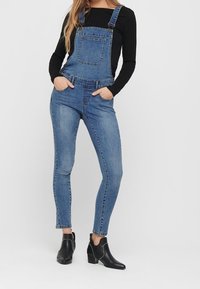 Blauwe spijkeroverall met verstelbare bandjes, voorzak en taps toelopende pasvorm; gecombineerd met zwarte enkellaarsjes met studs.