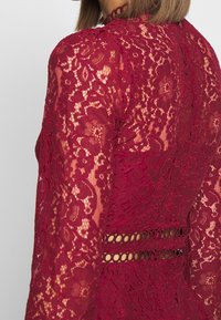 Bordeaux spetsklänning med långa ärmar, som har ett blommönster och cirkulära utskärningar vid midjan, vilket framhäver intrikata design detaljer.