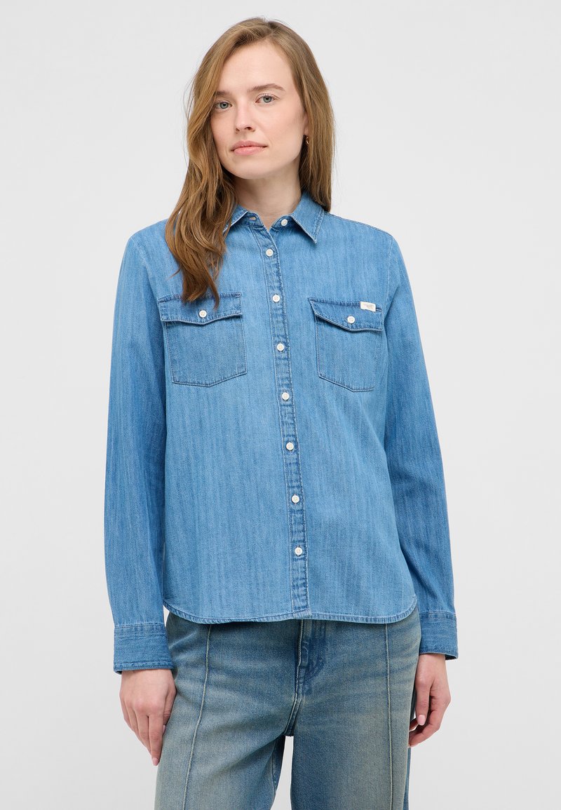 Kobieta w jasnoniebieskiej, dżinsowej koszuli na guziki z kieszeniami na piersi oraz w wyblakłych, niebieskich dżinsach, stojąca na jednolitym tle.