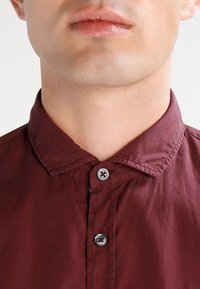 Detailní záběr dolní části obličeje a krku osoby, která má na sobě zapnutou vínovou košili s límečkem na jednoduchém pozadí.