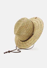 Kudottu olkihatun cowboy-hattu leveällä lierillä ja ruskealla säädettävällä leukahihnalla, vaalean valkoista taustaa vasten.