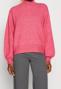 Pull en tricot rose à col roulé avec de longues manches bouffantes et des poignets et un ourlet côtelés. Porté avec un pantalon slim gris. Texture douce, design décontracté.