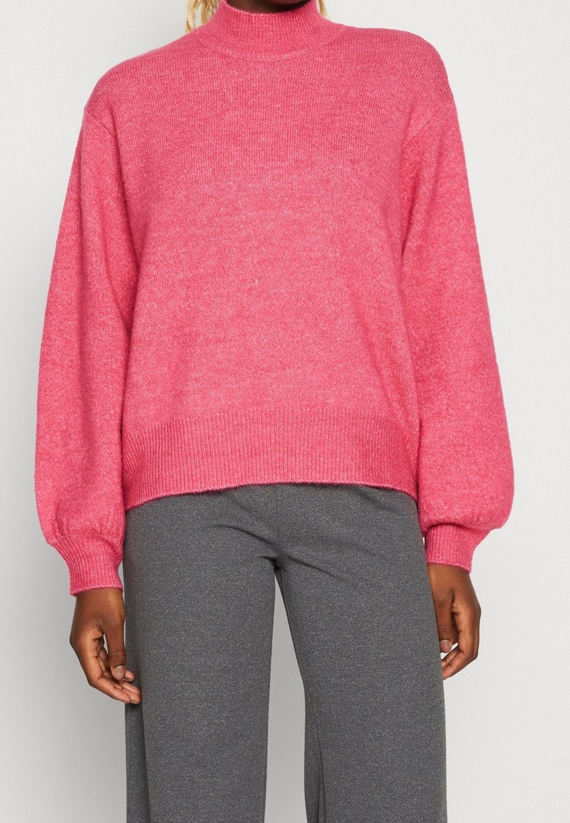 Pull en tricot rose à col roulé avec de longues manches bouffantes et des poignets et un ourlet côtelés. Porté avec un pantalon slim gris. Texture douce, design décontracté.
