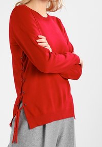 Maglione rosso a maniche lunghe con dettagli laterali in pizzo. Realizzato in un tessuto morbido con scollatura rotonda e polsini e orlo a costine.