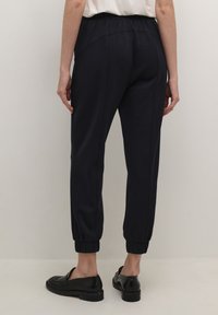 Marineblauwe joggerbroek met een ontspannen pasvorm, elastische tailleband en taps toelopende boorden. Glad textuur met zijnaad en zakken. G gedragen met zwarte instappers.