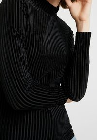 Schwarzes Langarmoberteil mit vertikaler Rippstruktur, hohem Kragen und elastischen Bündchen, das eine körpernahe Silhouette schafft.
