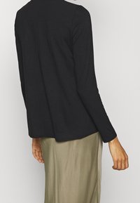 Svart långärmad topp gjord av mjukt tyg, med en avslappnad passform och rundad nederkant; i kombination med en ljus olivgrön kjol för kontrast.