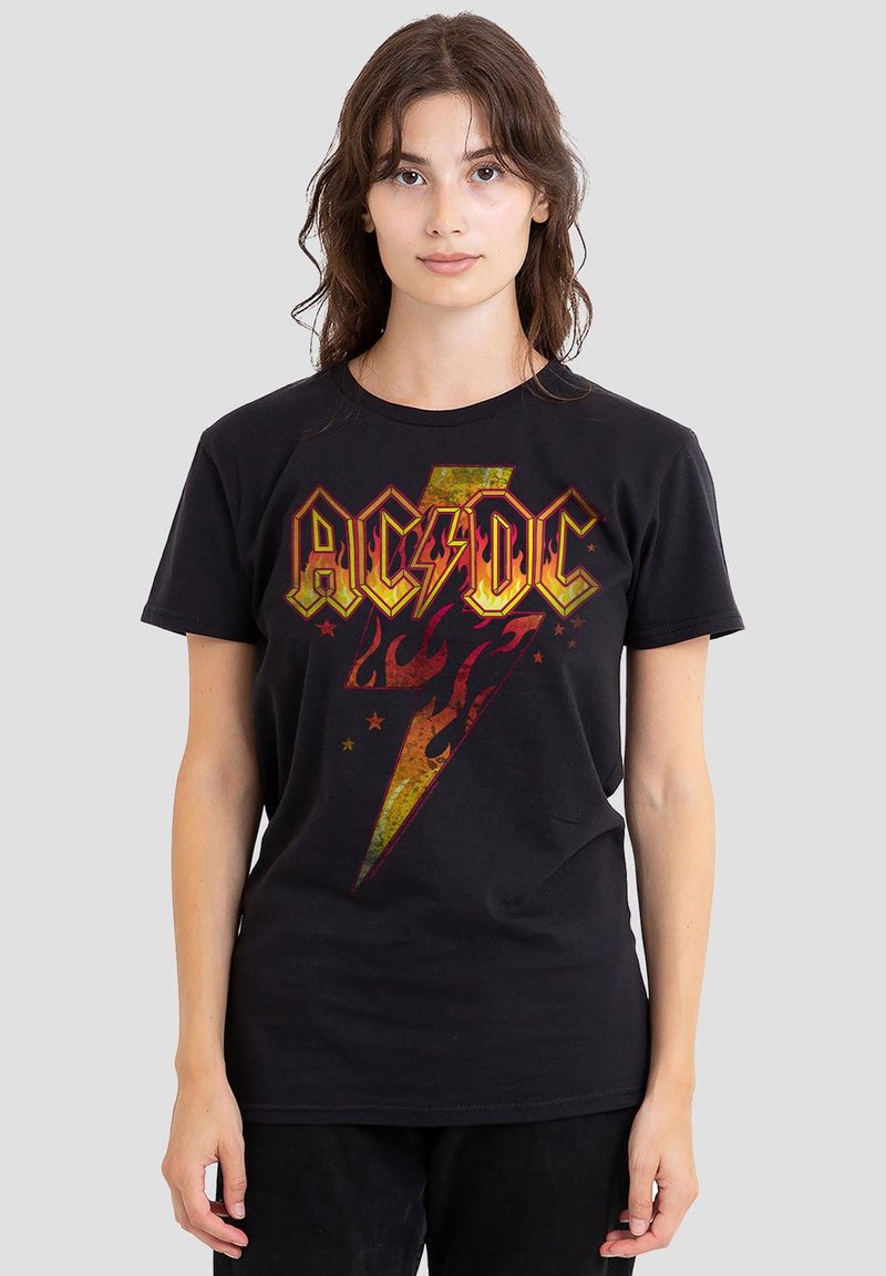 Junge Frau mit braunen Haaren trägt ein schwarzes AC/DC-Band-T-Shirt mit einem großen, vintage Blitz- und Flammendesign.